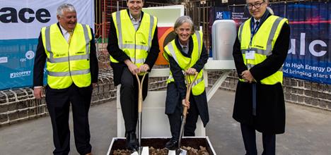 4 people in formal attire under high vis vests standing in a row smiling. The two in the middle hold shovels digging into soil.