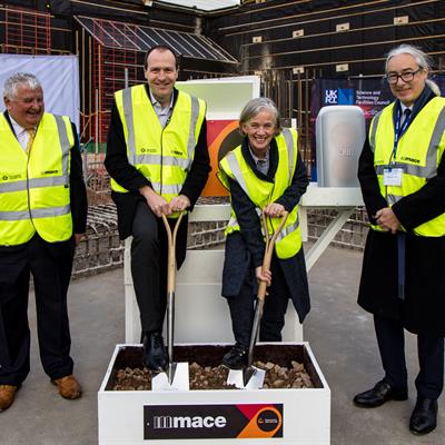 4 people in formal attire under high vis vests standing in a row smiling. The two in the middle hold shovels digging into soil.