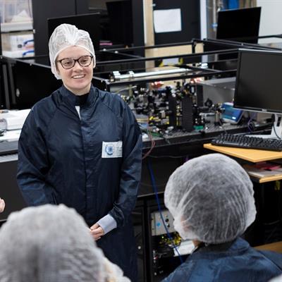 Danielle in a laser lab presenting to a group of young students, all in lab coats and hair nets. 