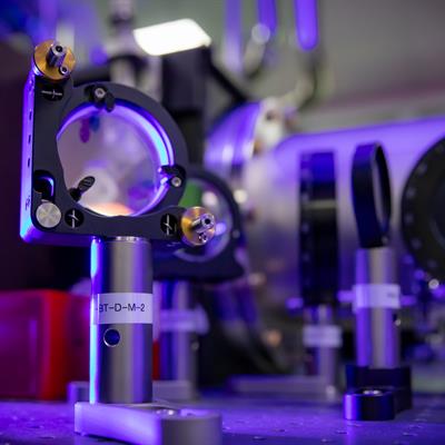 Close up of optics on a laser table. The lighting is deep purple and there is a pop of yellow on one of the optics.