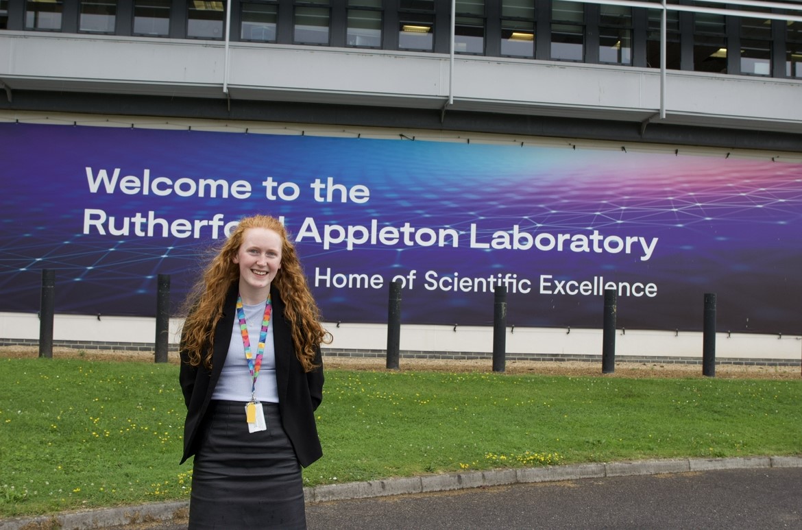 Ellen outside in front of a sign that reads "Welcome to the Rutherford Appleton Laboratory Home of Scientific Excellence"