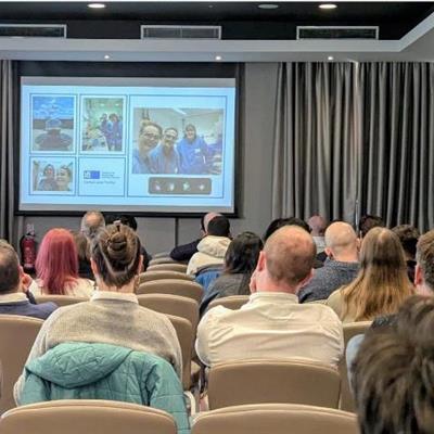 backs of peoples heads facing a presenter speaking with some slides projected on a wall.