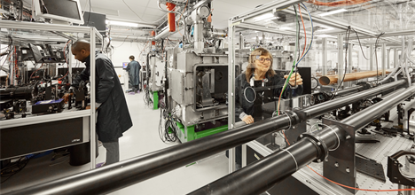 Bright and light lab full of chrome machinery. 3 scientists work in various places around the room in dark blue lab coats.