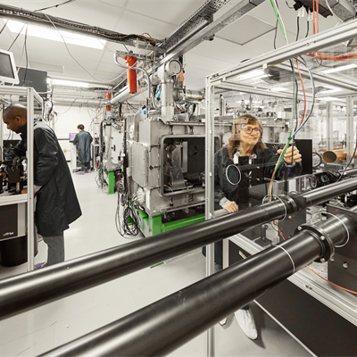 Bright and light lab full of chrome machinery. 3 scientists work in various places around the room in dark blue lab coats.