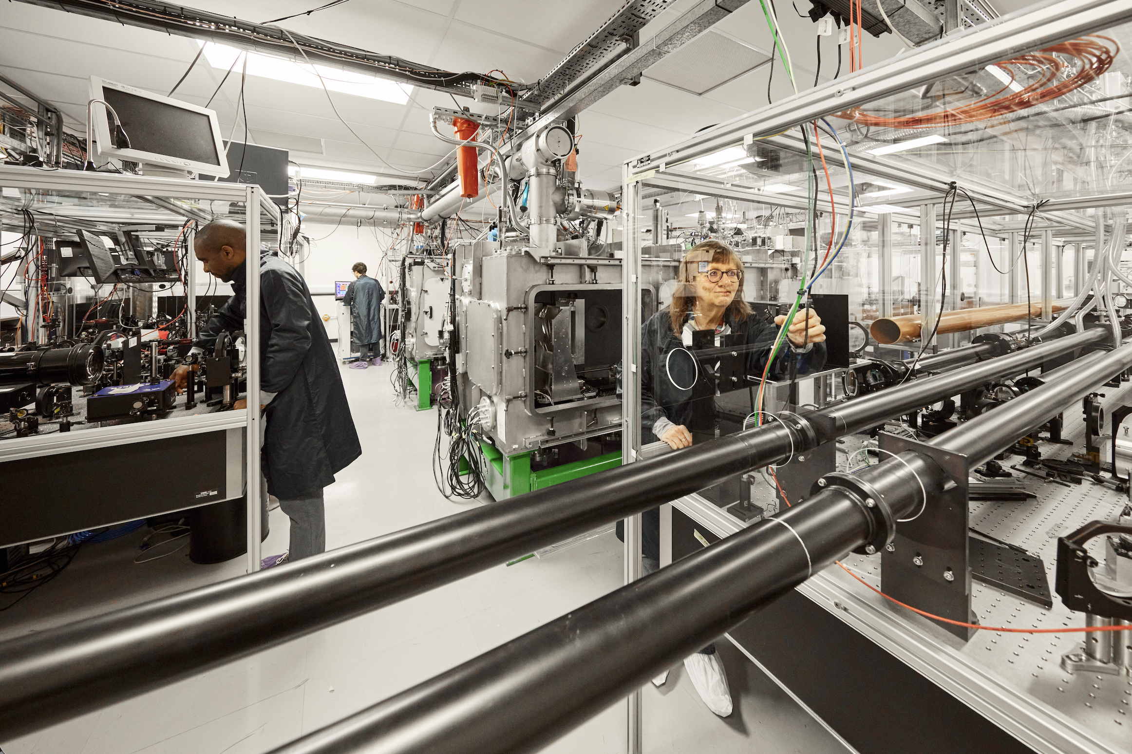 Bright and light lab full of chrome machinery. 3 scientists work in various places around the room in dark blue lab coats.