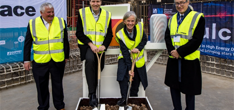 4 people in formal attire under high visibility vests stand smiling. One has a shovel and is digging into a box of dirt.