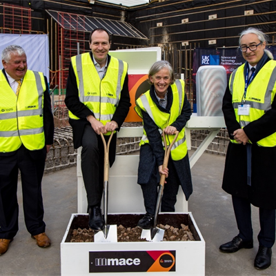 4 people in formal attire under high visibility vests stand smiling. One has a shovel and is digging into a box of dirt.