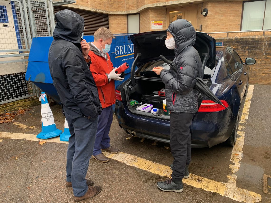 3 people in face masks stand beside an open car boot. One of them holds the SORS device.