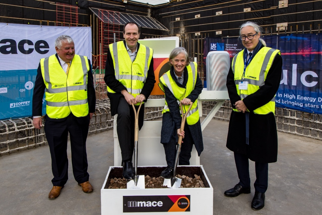 4 people in formal attire under high visibility vests stand smiling. One has a shovel and is digging into a box of dirt.